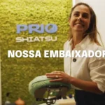 Carol Barcellos sorrindo em frente à parede verde com o logo da PRIO Shiatsu, segurando uma cadeira de massagem. Imagem destaca seu papel como embaixadora da PRIO, celebrando saúde, bem-estar e energia positiva no ambiente corporativo.