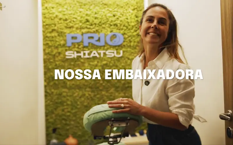 Carol Barcellos sorrindo em frente à parede verde com o logo da PRIO Shiatsu, segurando uma cadeira de massagem. Imagem destaca seu papel como embaixadora da PRIO, celebrando saúde, bem-estar e energia positiva no ambiente corporativo.
