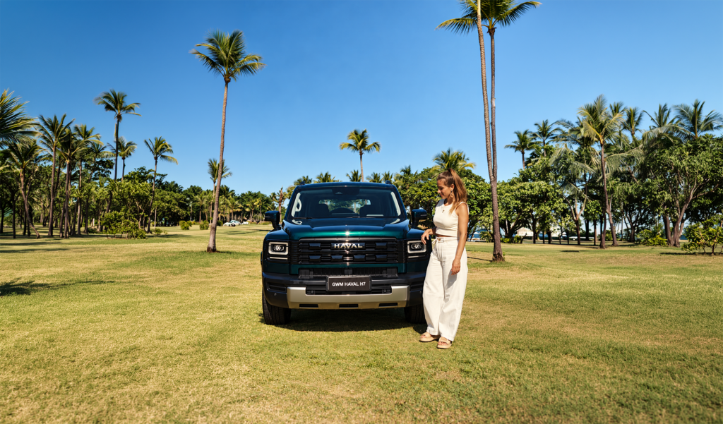 Carol Barcellos ao lado do GWM Haval H7 em um gramado com palmeiras ao fundo.