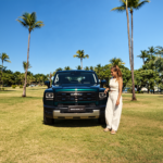 Carol Barcellos ao lado do GWM Haval H7 em um gramado com palmeiras ao fundo.